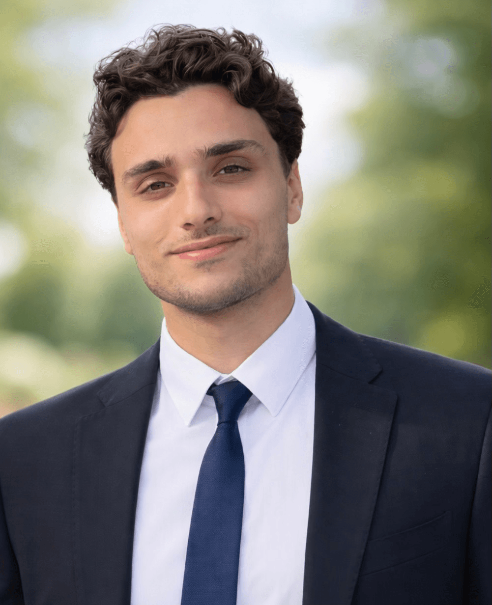 Professional headshot of Rayan Nehme wearing a dark suit and blue tie against a blurred outdoor background.
